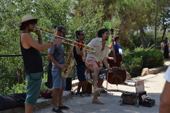 A group of street musicians are performing outdoors surrounded by greenery. The performers include a trombone player wearing a sun hat, a saxophonist, an upright bass player, and a guitarist who appears lively and animated. Equipment cases and a small box for donations are on the ground nearby.