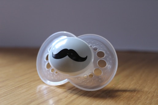 A pacifier with a humorous twist featuring a small black mustache design across the front. The pacifier is made of clear plastic with a smooth surface and multiple circular perforations. It is placed on a wooden surface with a soft shadow cast.