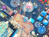 An aerial view of an amusement park featuring numerous rides, including a Ferris wheel, roller coasters, and colorful tents. The layout is vibrant and spacious, with pathways connecting different attractions. The area is bustling with activity, showcasing various structures and carnival rides laid out systematically.