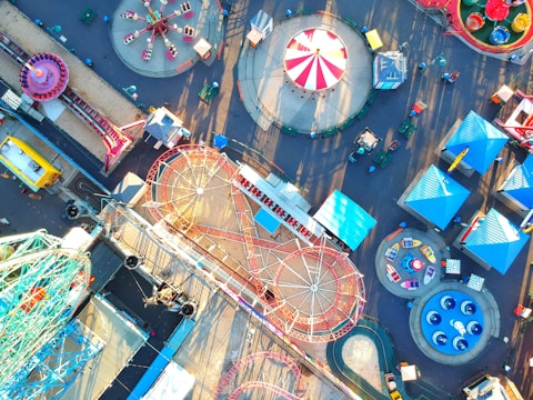 An aerial view of an amusement park featuring numerous rides, including a Ferris wheel, roller coasters, and colorful tents. The layout is vibrant and spacious, with pathways connecting different attractions. The area is bustling with activity, showcasing various structures and carnival rides laid out systematically.