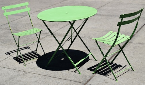 A green metal outdoor table and two matching folding chairs are placed on a concrete surface. The shadows cast by the midday sun create strong geometric patterns.