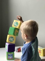 A young child is playing with colorful foam blocks, stacking them carefully in a vertical arrangement. The blocks feature various letters and illustrations, and the child's attention appears focused on maintaining the balance of the stack. A toy vehicle is visible on the table nearby.