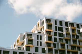 A modern apartment building with a geometric design, featuring numerous balconies and large windows. The structure is set against a clear blue sky with a few scattered clouds.