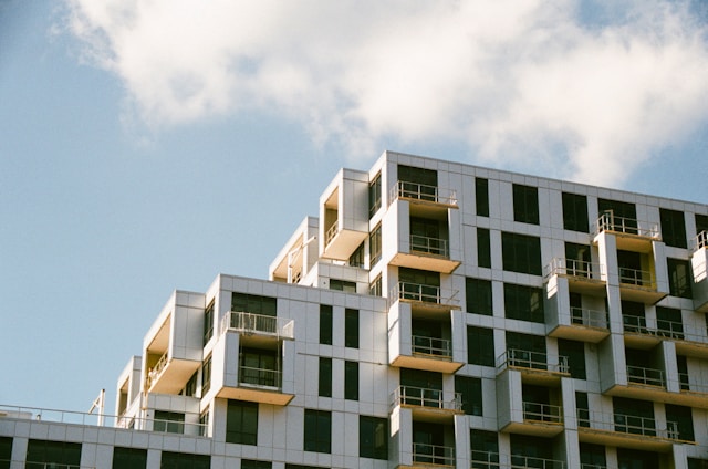 A modern apartment building with a geometric design, featuring numerous balconies and large windows. The structure is set against a clear blue sky with a few scattered clouds.