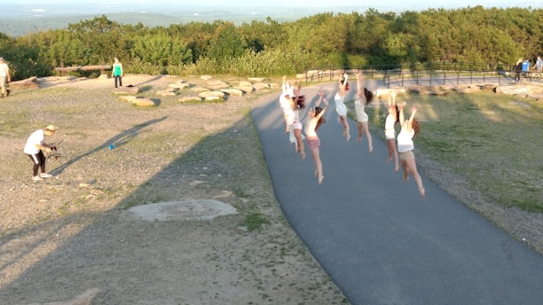 A group of people are energetically jumping in unison on a paved path surrounded by a natural, park-like setting. The participants, mostly dressed in light clothing, are in mid-air with their arms raised. In the background, lush greenery and rocks are visible. A person on the left is kneeling and appears to be photographing or filming the scene using a camera.