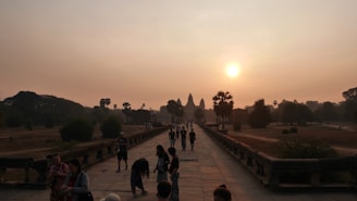 A crowd of people walks along a stone pathway toward a large, ancient temple in the distance. The temple features distinct spires and is set against a hazy sunrise. The atmosphere is calm with lush greenery and trees flanking the path.