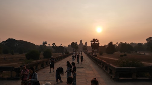 A crowd of people walks along a stone pathway toward a large, ancient temple in the distance. The temple features distinct spires and is set against a hazy sunrise. The atmosphere is calm with lush greenery and trees flanking the path.