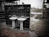 Two white ceramic sinks with pedestal bases are mounted on a dark tiled wall. The scene is outdoors with a brick pathway and a slightly overgrown grassy area nearby. There is a hose coiled on the ground and utility poles in the background, along with signs in a South Asian script.