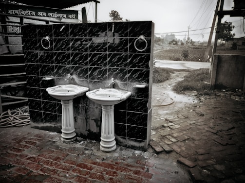 Two white ceramic sinks with pedestal bases are mounted on a dark tiled wall. The scene is outdoors with a brick pathway and a slightly overgrown grassy area nearby. There is a hose coiled on the ground and utility poles in the background, along with signs in a South Asian script.