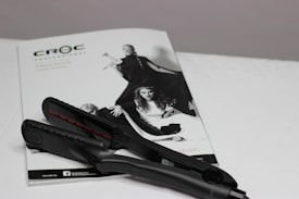 A black hair straightener is placed on top of a white magazine featuring fashion images. The magazine displays a monochromatic image of women in stylish clothing, with bold poses.