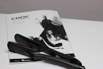 A black hair straightener is placed on top of a white magazine featuring fashion images. The magazine displays a monochromatic image of women in stylish clothing, with bold poses.