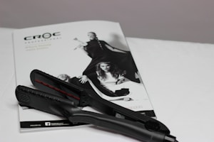 A black hair straightener is placed on top of a white magazine featuring fashion images. The magazine displays a monochromatic image of women in stylish clothing, with bold poses.