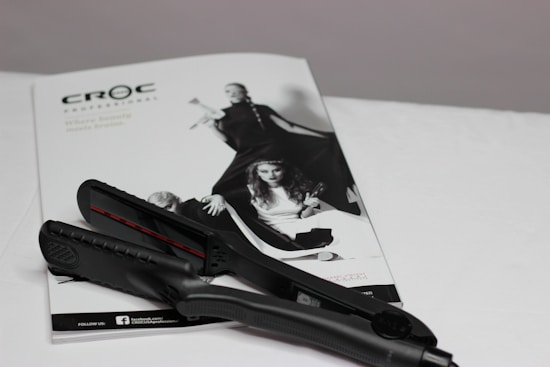 A black hair straightener is placed on top of a white magazine featuring fashion images. The magazine displays a monochromatic image of women in stylish clothing, with bold poses.