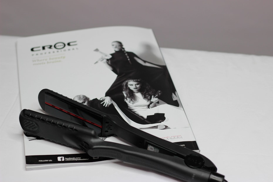 A black hair straightener is placed on top of a white magazine featuring fashion images. The magazine displays a monochromatic image of women in stylish clothing, with bold poses.