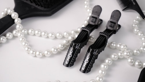 A flat lay arrangement featuring black hair styling clips branded 'Croc', a black paddle brush, a black comb, and a white pearl necklace. The background is a soft white fabric, creating a contrast with the black tools and the shimmering pearls.