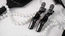 A flat lay arrangement featuring black hair styling clips branded 'Croc', a black paddle brush, a black comb, and a white pearl necklace. The background is a soft white fabric, creating a contrast with the black tools and the shimmering pearls.