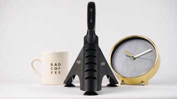 A white ceramic mug printed with 'BAD COFFEE' stands next to a sleek black rocket-shaped object. Behind them is a stylish gold and white clock with marble texture on its face.