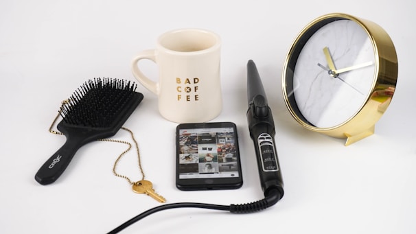 A composition of everyday items including a black hairbrush, a mug with 'BAD COFFEE' printed on it, a smartphone displaying a grid of images, a black hair curling wand, a golden key on a chain, and a gold-rimmed clock with a marble pattern face. The objects are arranged neatly on a white surface.