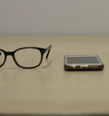 A pair of black-rimmed glasses, a smartphone, and a smartwatch are arranged side by side on a beige surface. The items suggest a modern, tech-oriented lifestyle.