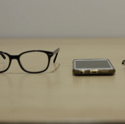 A pair of black-rimmed glasses, a smartphone, and a smartwatch are arranged side by side on a beige surface. The items suggest a modern, tech-oriented lifestyle.
