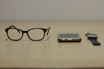 A pair of black-rimmed glasses, a smartphone, and a smartwatch are arranged side by side on a beige surface. The items suggest a modern, tech-oriented lifestyle.