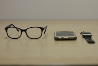 A pair of black-rimmed glasses, a smartphone, and a smartwatch are arranged side by side on a beige surface. The items suggest a modern, tech-oriented lifestyle.