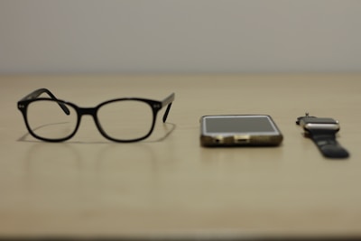 A pair of black-rimmed glasses, a smartphone, and a smartwatch are arranged side by side on a beige surface. The items suggest a modern, tech-oriented lifestyle.