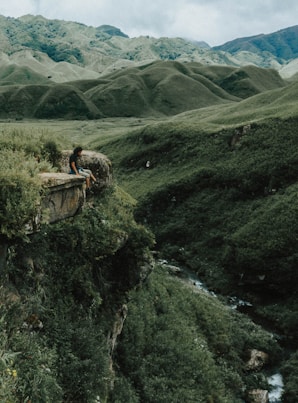 man sitting on brown rock on cliff with distance to mountains