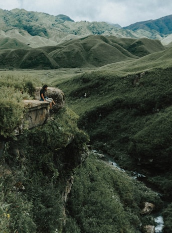 man sitting on brown rock on cliff with distance to mountains