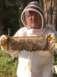 A person wearing a beekeeping suit, including a protective hat and veil, is holding a frame covered in bees. The background includes trees and greenery, suggesting an outdoor apiary setting. The person's face is visible behind glasses, and they appear to be smiling.