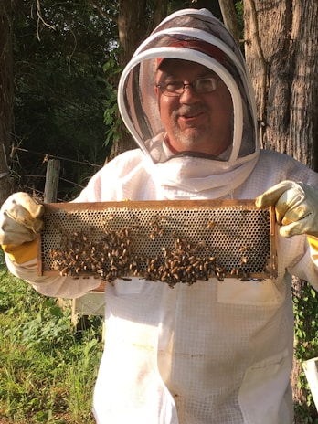 A person wearing a beekeeping suit, including a protective hat and veil, is holding a frame covered in bees. The background includes trees and greenery, suggesting an outdoor apiary setting. The person's face is visible behind glasses, and they appear to be smiling.
