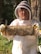 A person wearing a beekeeping suit, including a protective hat and veil, is holding a frame covered in bees. The background includes trees and greenery, suggesting an outdoor apiary setting. The person's face is visible behind glasses, and they appear to be smiling.