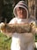 A person wearing a beekeeping suit, including a protective hat and veil, is holding a frame covered in bees. The background includes trees and greenery, suggesting an outdoor apiary setting. The person's face is visible behind glasses, and they appear to be smiling.
