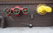Three red valves with green wheels are mounted on a brown brick wall. To the right, a yellow standpipe with a chain is also mounted on the wall. Below, there are two outlets and a black cylindrical object on the concrete floor. A rusted pipe extends from the left side.