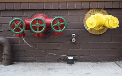 Three red valves with green wheels are mounted on a brown brick wall. To the right, a yellow standpipe with a chain is also mounted on the wall. Below, there are two outlets and a black cylindrical object on the concrete floor. A rusted pipe extends from the left side.