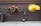 Three red valves with green wheels are mounted on a brown brick wall. To the right, a yellow standpipe with a chain is also mounted on the wall. Below, there are two outlets and a black cylindrical object on the concrete floor. A rusted pipe extends from the left side.
