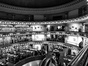 A multi-level bookstore with ornate architectural details including decorative columns and arches. Shelves filled with books line the walls of each level, and people are browsing and reading. An escalator is visible in the foreground, connecting the different floors.