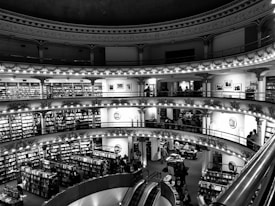 A multi-level bookstore with ornate architectural details including decorative columns and arches. Shelves filled with books line the walls of each level, and people are browsing and reading. An escalator is visible in the foreground, connecting the different floors.