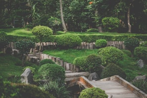 A serene garden landscape characterized by well-manicured bushes and trees, arranged in a harmonious manner with natural wooden elements marking pathways. The lush greenery is abundant, contributing to the tranquil atmosphere. Stone elements complement the natural aesthetic, and the area is surrounded by dense woods.