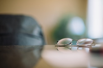 A pair of eyeglasses resting on a reflective surface with a blurred background, suggesting a tranquil and soft setting.