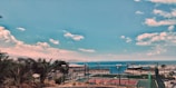 An outdoor sports court complex with tennis courts, surrounded by fences. Beyond the courts lies a coastal view with a calm sea and a distant city skyline. Lush palm trees and shrubbery are visible in the foreground, and the sky is clear with some scattered clouds.