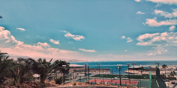 An outdoor sports court complex with tennis courts, surrounded by fences. Beyond the courts lies a coastal view with a calm sea and a distant city skyline. Lush palm trees and shrubbery are visible in the foreground, and the sky is clear with some scattered clouds.