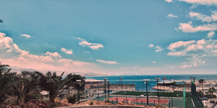 An outdoor sports court complex with tennis courts, surrounded by fences. Beyond the courts lies a coastal view with a calm sea and a distant city skyline. Lush palm trees and shrubbery are visible in the foreground, and the sky is clear with some scattered clouds.