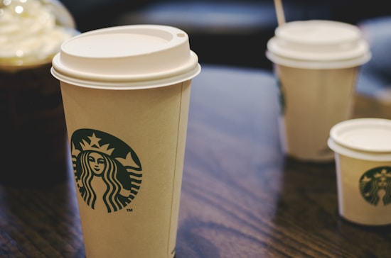 A close-up of several paper coffee cups with lids placed on a dark wooden table. The cups prominently feature the Starbucks logo and vary in size, with one larger cup in the foreground and smaller cups in the background. The lighting is soft, enhancing the warm tones of the scene.