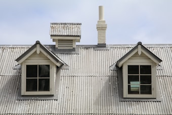 A weathered corrugated metal roof with two small dormer windows and a chimney. The windows have gray frames and are symmetrically placed. The roof exhibits signs of age and peeling paint.
