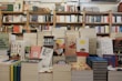 A bookstore with shelves filled with a variety of books arranged neatly. There is a table in the foreground stacked with several different books with colorful covers, featuring notable authors and titles. Books are stacked both vertically and horizontally, showcasing different genres.