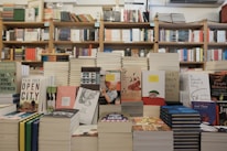A bookstore with shelves filled with a variety of books arranged neatly. There is a table in the foreground stacked with several different books with colorful covers, featuring notable authors and titles. Books are stacked both vertically and horizontally, showcasing different genres.