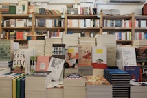 A bookstore with shelves filled with a variety of books arranged neatly. There is a table in the foreground stacked with several different books with colorful covers, featuring notable authors and titles. Books are stacked both vertically and horizontally, showcasing different genres.