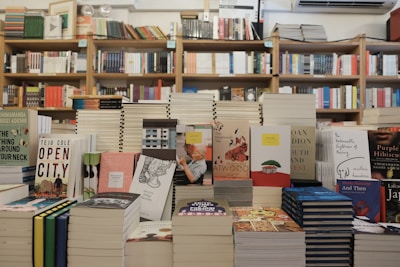 A bookstore with shelves filled with a variety of books arranged neatly. There is a table in the foreground stacked with several different books with colorful covers, featuring notable authors and titles. Books are stacked both vertically and horizontally, showcasing different genres.