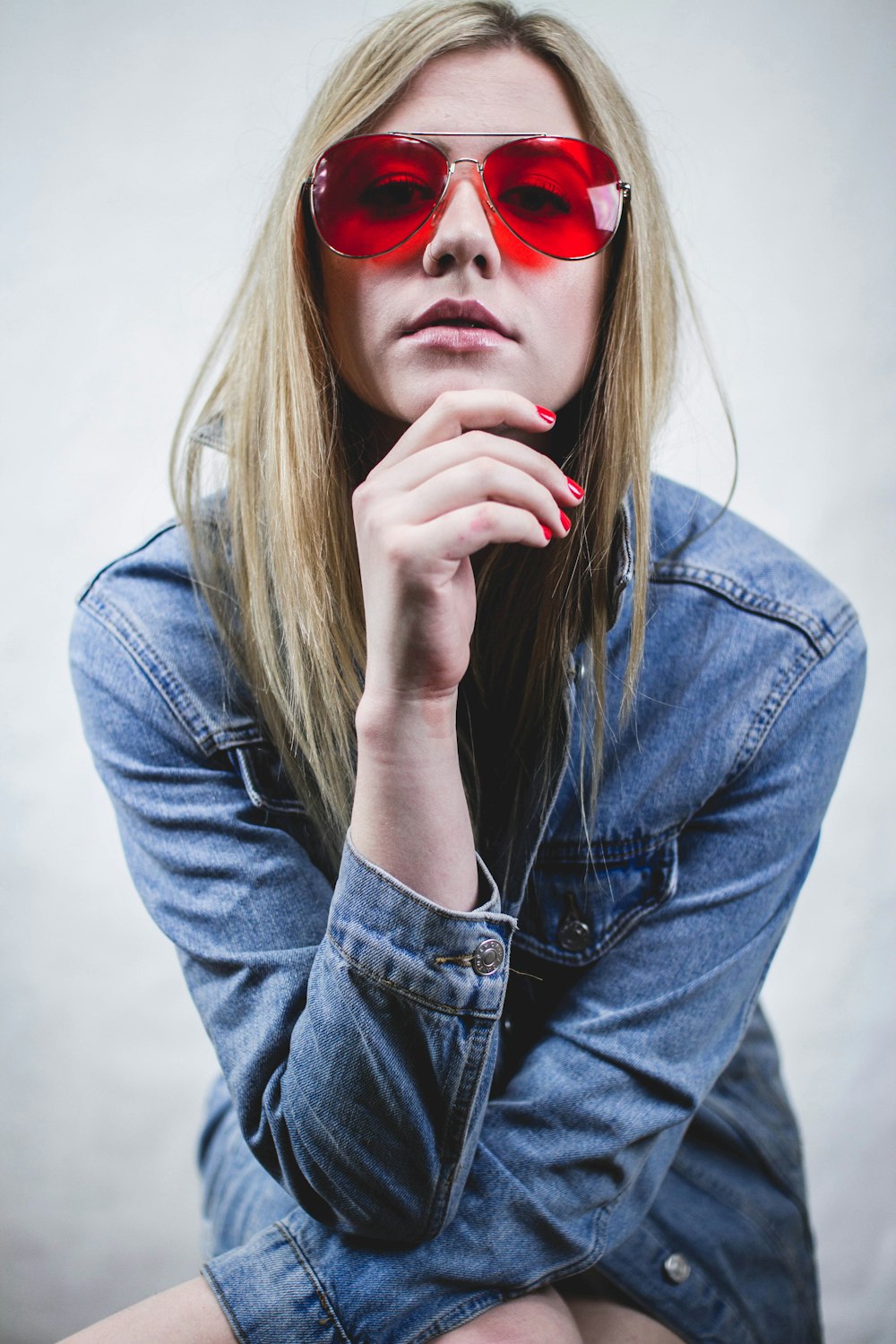 Donna in giacca di jeans blu che indossa occhiali da sole rossi e manicure rossa con le mani sul mento